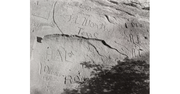 A black-and-white, tightly focused photograph of a large rock with carved inscriptions, including various initials and the words "Ft. Worth, Texas."