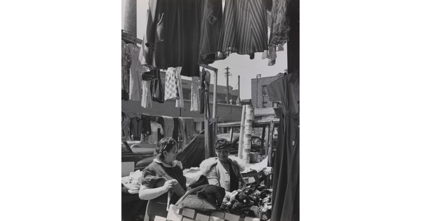 A black-and-white photograph of two light- to medium-skinned women smiling at each other in an open-air market, surrounded by clothing and boxes.