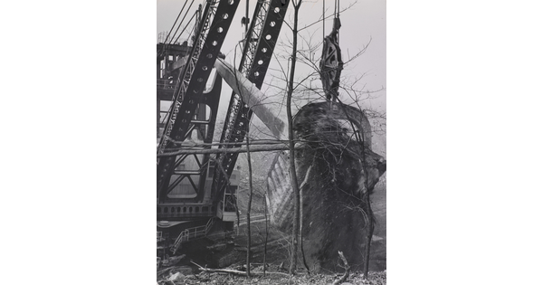 A black-and-white photograph of a massive digging machine taking a huge chunk of earth out of the ground with spindly trees in the foreground.