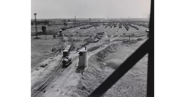 A black-and-white photograph of several train cars on tracks that lead to a train yard filled with more cars in the distance.