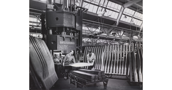 A black-and-white photograph of two men in a factory holding an airplane propeller surrounded by racks of propellers.