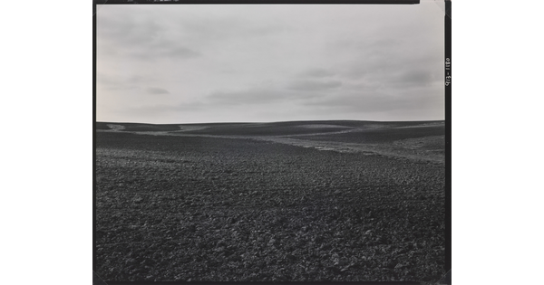 A black-and-white photograph of a dirt landscape with subtle hills under a cloudy sky.