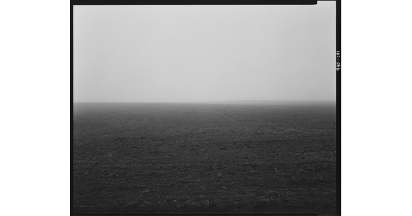 A black-and-white photograph of a dirt landscape with a hazy horizon and a subtle hill in the distance.