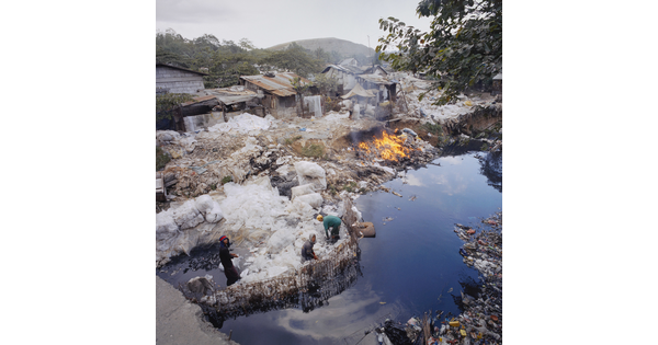 A color photograph of people sorting through a garbage dump next to water, several shacks in the background, and a fire going next to the water.