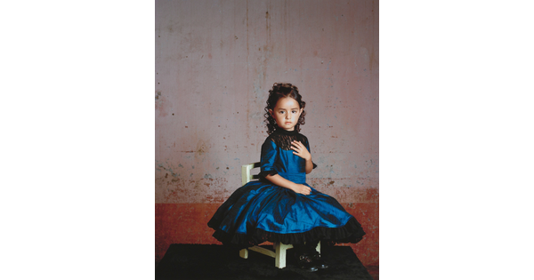 A color photograph of a young girl in a formal blue dress posed on a white chair.