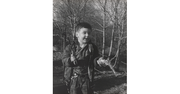 A black-and-white photograph of a White boy playing with two toy revolvers in a wooded setting.