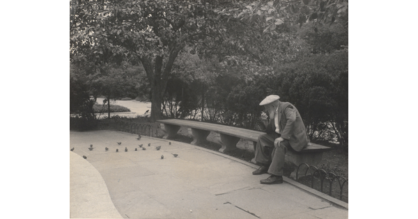 A black-and-white photograph of an older White man with a beard sitting on a stone bench in a park.