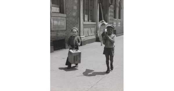 A black-and-white photograph of a White child banging a box like a drum and a Black child waving a flag of crumpled paper as they march on a city sidewalk.
