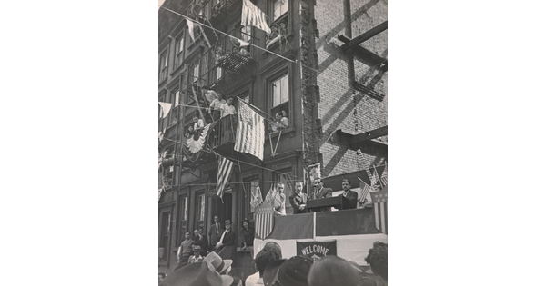 A black-and-white photograph of a man making a speech on a platform with people watching and American flags hanging from apartment windows above.