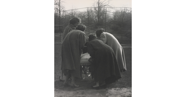 A black-and-white photograph of four women, backs to the viewer, bending down to look in a baby buggy.