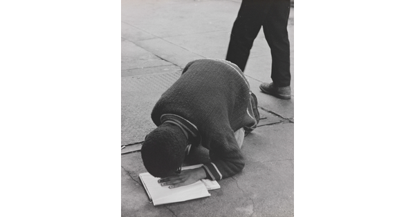 A black-and-white photograph of a Black boy hunched over his knees on pavement, his hand on a book and face close to his hand.