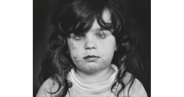 A black-and-white photograph of a White, toddler girl with dark, curly hair and dirt on her cheeks looking directly at the viewer.