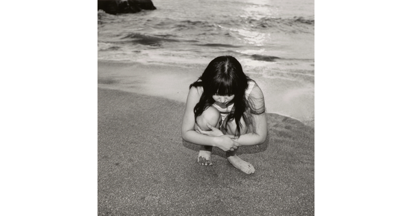 A black-and-white photograph of a young White girl with long dark hair crouched on a shoreline looking at the sand.