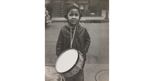 A black-and-white photograph of a warmly dressed young boy with medium-toned skin wearing a drum from his neck and looking directly into camera.