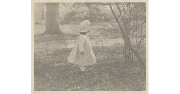 A black-and-white photograph of a young child in a white dress and hat standing in a field of grass and facing away from the camera.