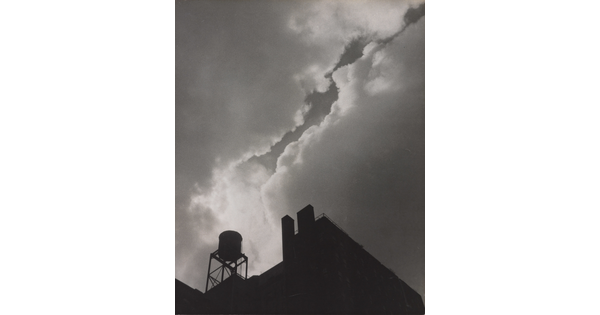 A black-and-white photograph taken from the ground looking up of silhouettes of industrial buildings and a water tower against a cloudy sky.