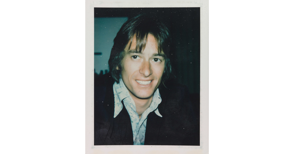 An informal color photograph of a smiling White man from the shoulders up with longish hair, wearing an open collar shirt and dark jacket.