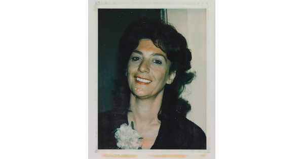 An informal color photograph of a smiling White woman from the shoulders up with dark, curly hair, red lipstick, and a white carnation on her black shirt.