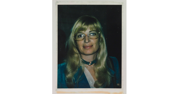 An informal color photograph of a White woman with blonde hair and tortoise-shell glasses wearing a blue suit and necklace.