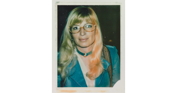 An informal color photograph of a White woman with blonde hair and tortoise-shell glasses wearing a blue suit and necklace.