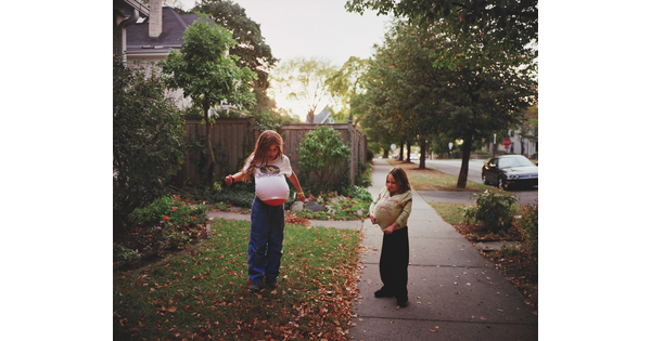 A color photograph of two young White girls playing in a front yard with balloons under their shirts mimicking pregnancy.