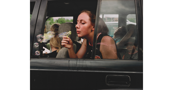 A color photograph of a White teenage girl blowing bubbles out of a backseat car window.