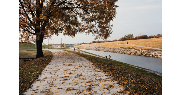 A color photograph of a late fall landscape with a river disappearing into the distance and people running on well-groomed paths.