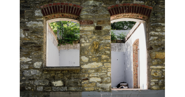A color photograph of two brick arches, one a window the other a door, in a stone wall, looking into an empty, white room with no roof and green vegetation overhead.