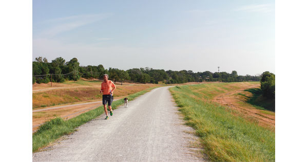 A color photograph of a middle-aged White man and an unleashed dog running toward the viewer on a paved path.