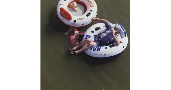 A color photograph taken from above of a White man reclining in an innertube and a White woman in pink swimsuit soaking in the water next to an innertube.