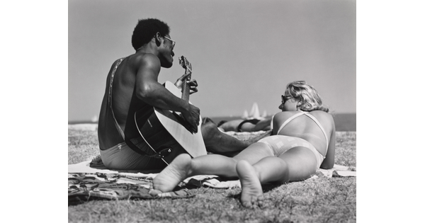 A black-and-white photograph of a Black man playing guitar on a beach blanket next to a White woman lying on her stomach with her feet toward the camera.
