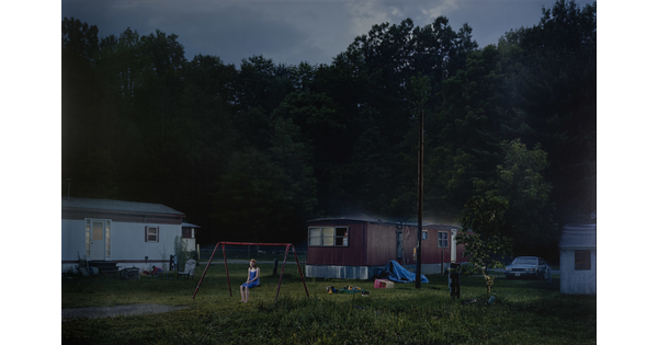 A color photograph of a White teenage girl sitting on a swing set in front of a mobile home park.