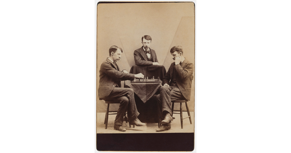 A sepia-toned photograph of the same White man with a moustache wearing a suit seated in three different positions around a chess board.
