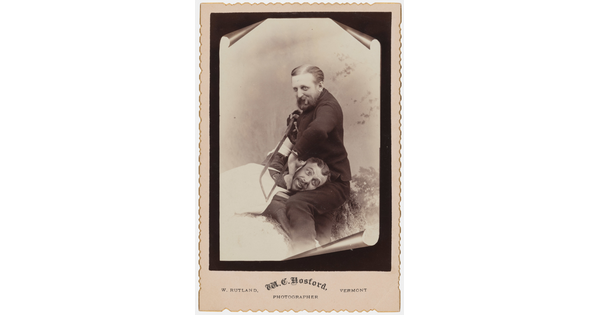 A sepia-toned studio photograph of a seated White man holding a saw against the neck of another White man whose head is resting on his lap.