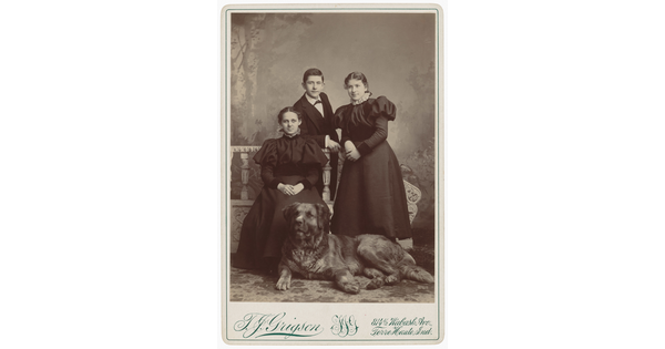 A black-and-white studio portrait photograph of three young adults, two White women and one White man, posing behind a large dog.