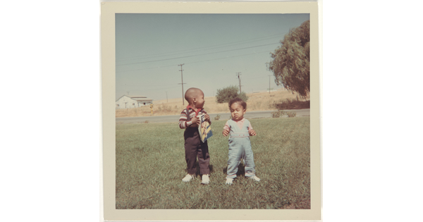 A color photograph of two Black toddlers standing in a grassy yard.
