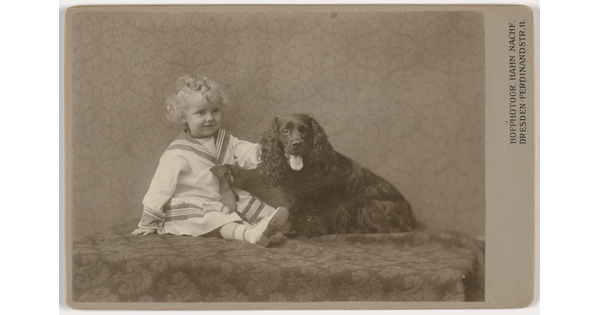 A sepia-toned photograph of a young White child with blond ringlets wearing a sailor dress sitting next to an Irish setter-like dog.