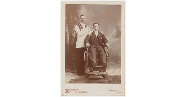 A sepia-toned photograph of two White men in Victorian clothing, one seated in a barber's chair, the other standing behind the chair holding scissors.
