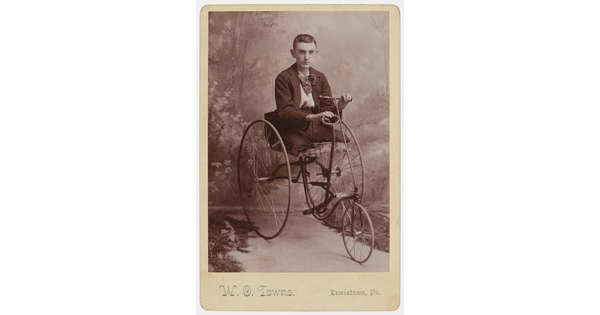 A sepia-toned studio photograph of teenage White boy with no legs, seated in a Victorian tricycle-style wheelchair.