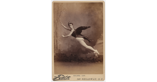 A sepia-toned studio photograph of a White woman, black wings on her back, arms extended, suspended in the air as if flying.