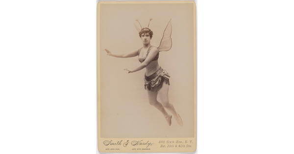 A sepia-toned studio photograph of a White woman dressed as a fairy, including wings and antennae, suspended in the air against a white background.