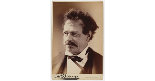 A sepia-toned studio portrait photograph of a middle-aged White man with unkempt hair, a mustache, wearing a black jacket, white shirt with a high collar, and bow tie.