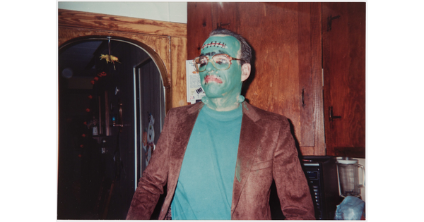 A color photograph of a man wearing green face paint and a Frankenstein costume standing in what looks like a kitchen.