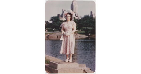 A color photograph of a White woman wearing a decorative sunhat and a floral dress standing on a concrete block beside a pond with tall buildings in the background.