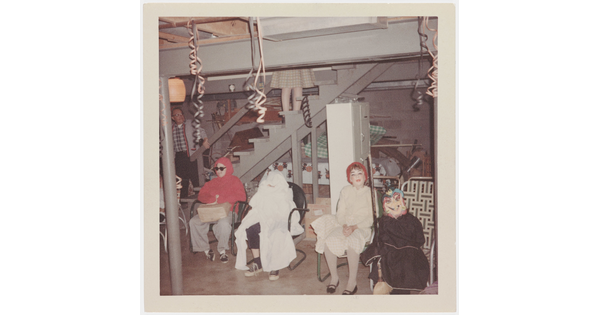 A faded color photograph of four children in costumes sitting in chairs at the foot of basement stairs.
