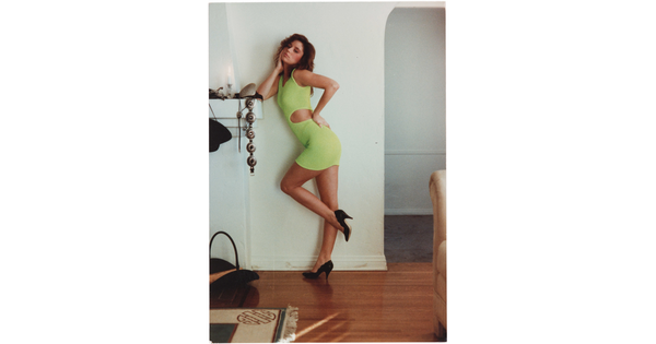 A color photograph of a White woman in a bright green dress and black heels, one hand on her hip the other elbow on a fireplace mantel, striking a pose for the camera.