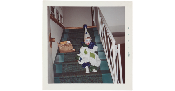 A color snapshot of a toddler in a blue, green, and white clown costume sitting on stairs inside a house.