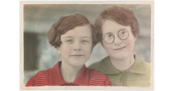 A color photograph of two White children with short auburn hair, the child on the right wears glasses, leaning their heads toward each other.