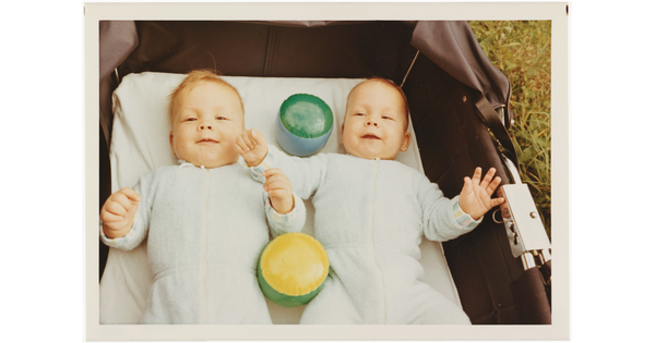 A color photograph of two White babies smiling and lying on their backs with toys in a baby carriage that is sitting in grass.