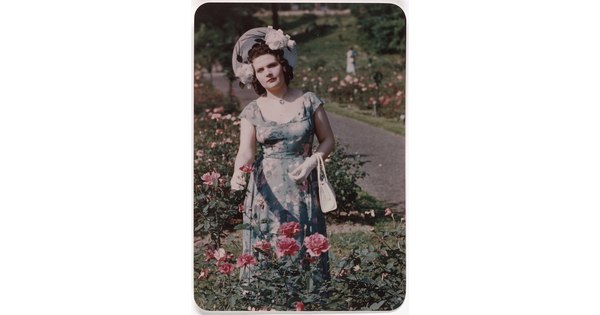 A color photograph of a White woman wearing a sunhat, floral dress, and white gloves standing in a rose garden.